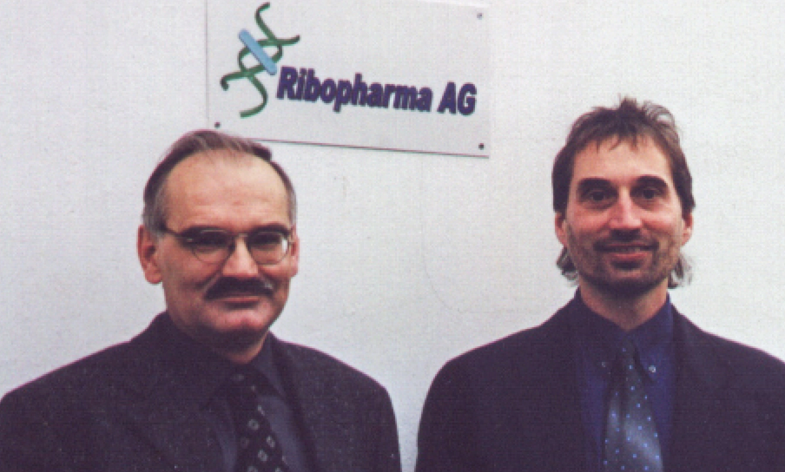 Two men in formal attire stand in front of a wall-mounted sign that reads &quot;Ribopharma AG.&quot; The man on the left wears glasses and has a mustache, while the man on the right has a short beard and longer hair. Both are smiling slightly. The background is plain, drawing attention to the individuals and the company sign.