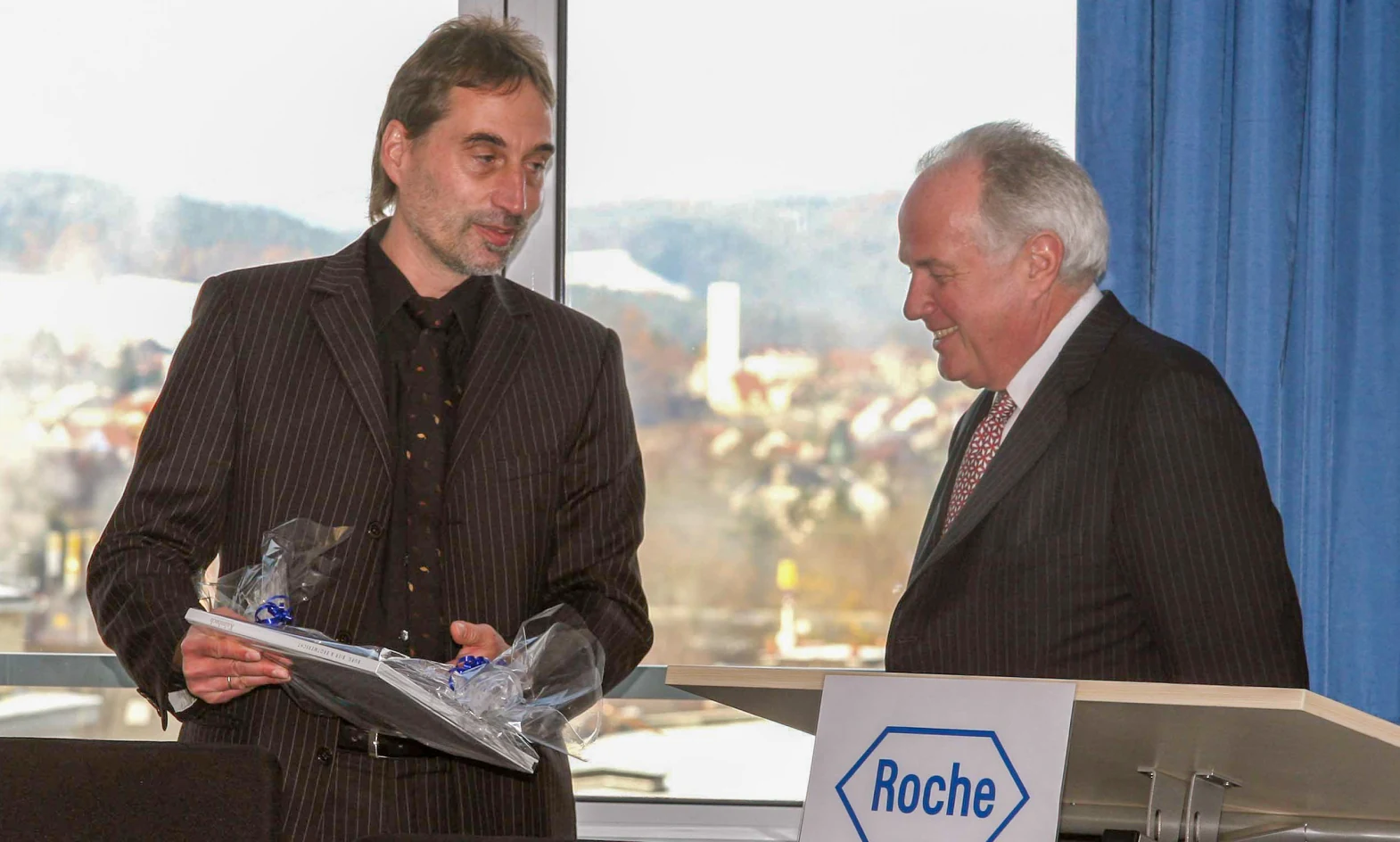 Two men in suits are smiling and conversing at a podium with a &quot;Roche&quot; logo, as one presents a wrapped gift or award to the other.