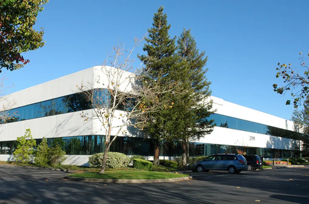 Modern two-story office building with large reflective glass windows surrounded by trees and a parking lot under a clear blue sky.