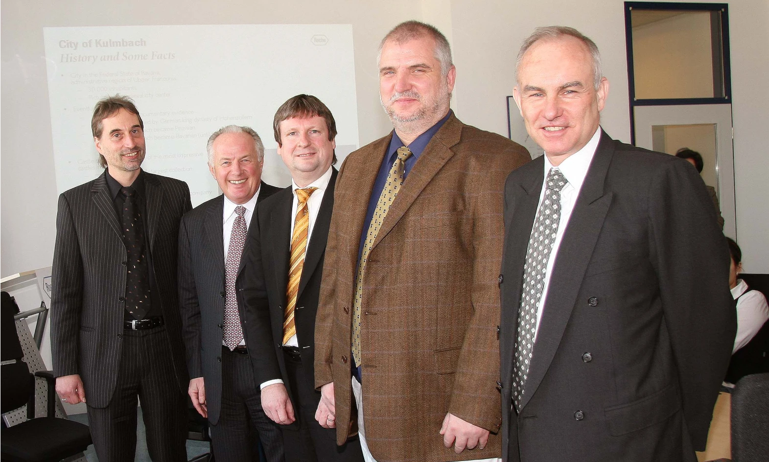 Five professionally dressed men standing in a row and smiling at the camera during a formal event or business meeting. A presentation slide titled &#039;City of Kulmbach: History and Some Facts&#039; is projected in the background.