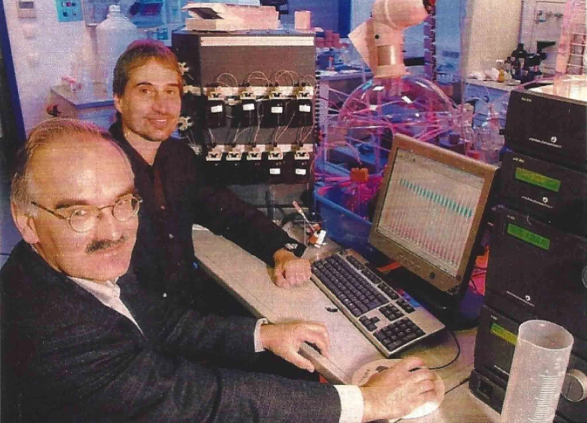 Two scientists working at a computer station in a laboratory, with laboratory automation equipment and data analysis on the monitor.